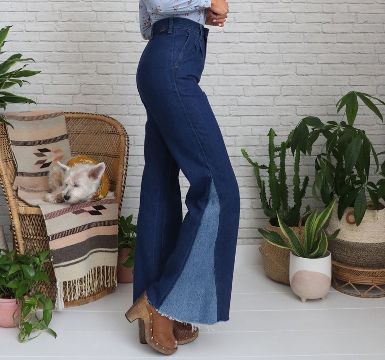 Pode incluir: Uma mulher vestindo um par de cal&ccedil;as jeans de sino azul de denim com um painel azul contrastante na parte inferior de cada perna. As cal&ccedil;as jeans t&ecirc;m cintura alta e um caimento relaxado.