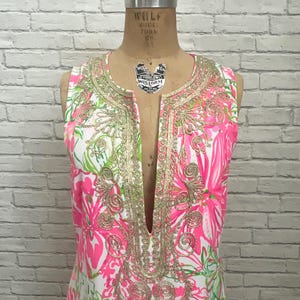 May include: A sleeveless tunic dress featuring a vibrant pink, white, and green floral print. The garment has a deep V-neckline adorned with intricate gold metallic embroidery in a swirling pattern that extends down the front placket.