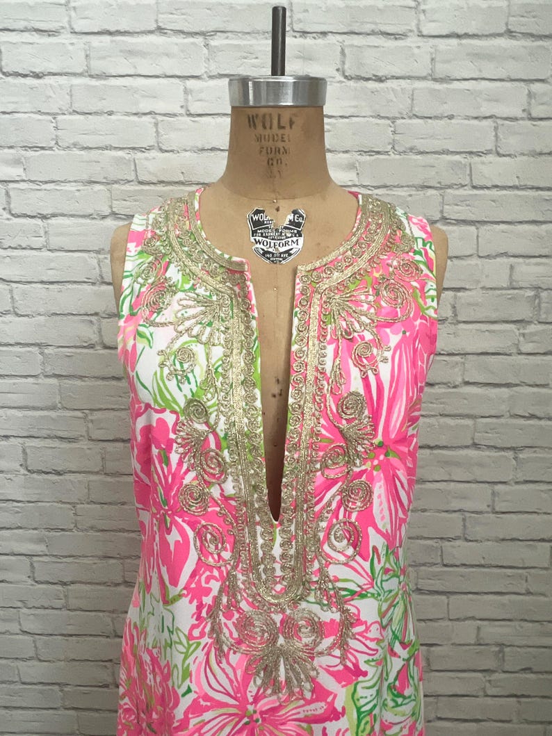 May include: A sleeveless tunic dress featuring a vibrant pink, white, and green floral print. The garment has a deep V-neckline adorned with intricate gold metallic embroidery in a swirling pattern that extends down the front placket.