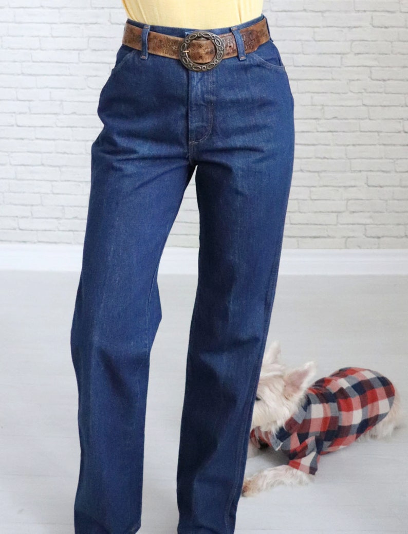 May include: A pair of dark blue denim jeans with a brown leather belt featuring a large metal buckle. The jeans have a high waist and a straight leg.