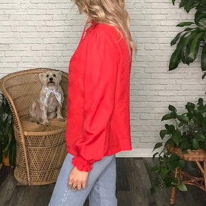 May include: A red long-sleeved blouse with a ruffled neckline and long sleeves. The blouse is made of a lightweight fabric and has a relaxed fit.