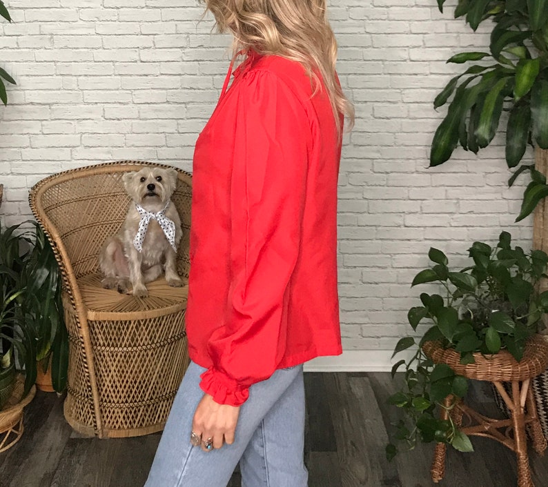 May include: A red long-sleeved blouse with a ruffled neckline and long sleeves. The blouse is made of a lightweight fabric and has a relaxed fit.