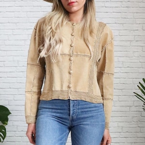 May include: A tan suede jacket with a crocheted trim and button closure. The jacket has a patchwork design and is cropped.
