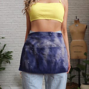 May include: A denim apron with a blue tie-dye effect and white paint splatters. The apron has a large pocket and is worn over blue and white striped pants. A yellow crop top is also visible.