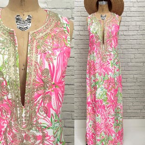 May include: A sleeveless floor-length maxi dress featuring a vibrant pink, white, and lime green floral print. The garment has a deep notched neckline adorned with intricate gold metallic embroidery and is paired with a wide-brimmed straw hat.