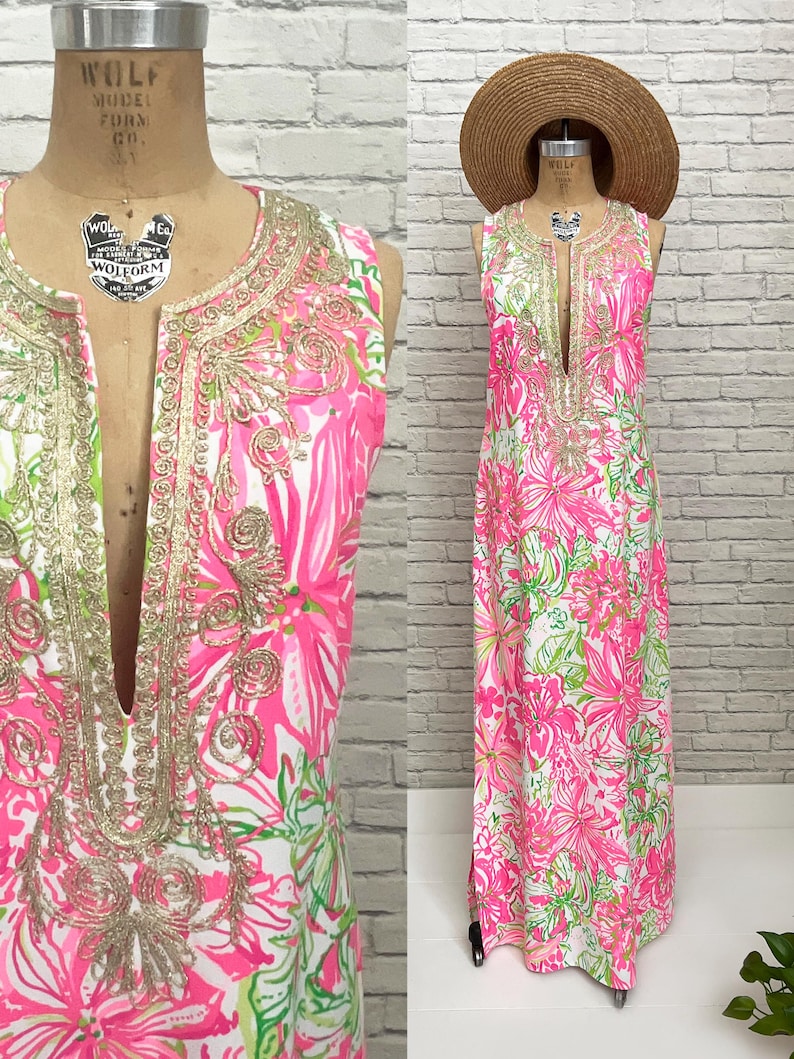 May include: A sleeveless floor-length maxi dress featuring a vibrant pink, white, and lime green floral print. The garment has a deep notched neckline adorned with intricate gold metallic embroidery and is paired with a wide-brimmed straw hat.