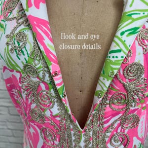 May include: A white sleeveless garment featuring a vibrant pink and green floral pattern with metallic gold embroidery along the collar and neckline. The front includes a series of metal hook and eye closures with the text Hook and eye closure details.