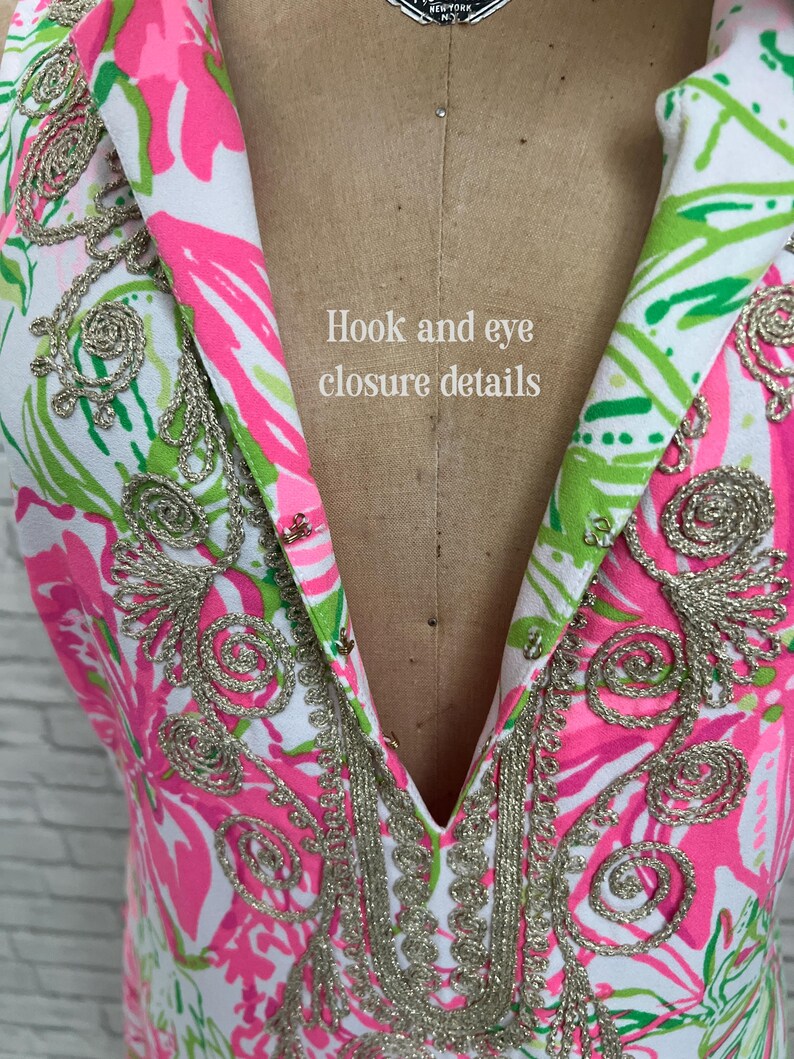 May include: A white sleeveless garment featuring a vibrant pink and green floral pattern with metallic gold embroidery along the collar and neckline. The front includes a series of metal hook and eye closures with the text Hook and eye closure details.