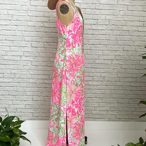 May include: A sleeveless floor-length dress featuring a vibrant pink, white, and green floral pattern with a side slit, displayed on a mannequin wearing a straw hat. The dress has a fitted bodice and a straight silhouette that extends to the floor.