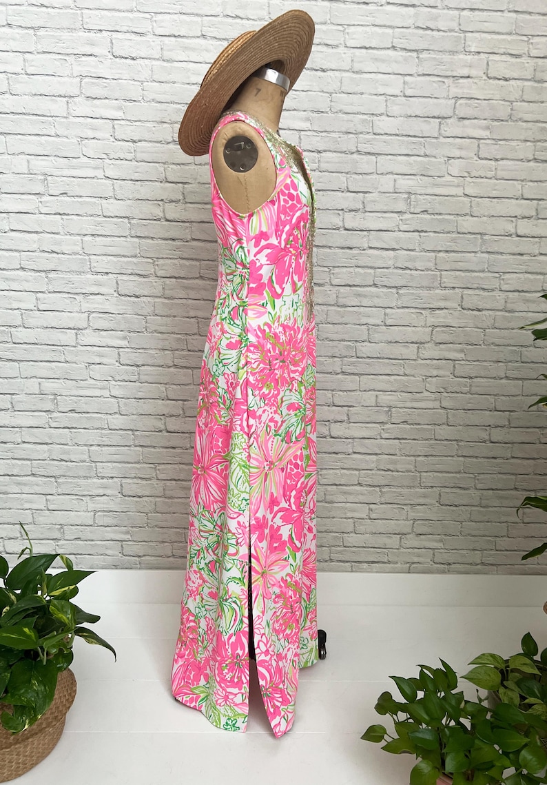 May include: A sleeveless floor-length dress featuring a vibrant pink, white, and green floral pattern with a side slit, displayed on a mannequin wearing a straw hat. The dress has a fitted bodice and a straight silhouette that extends to the floor.