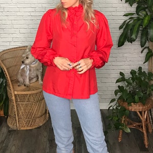 May include: A red long-sleeved blouse with a ruffled collar and button-up front. The blouse has puffed sleeves with ruffles at the cuffs.