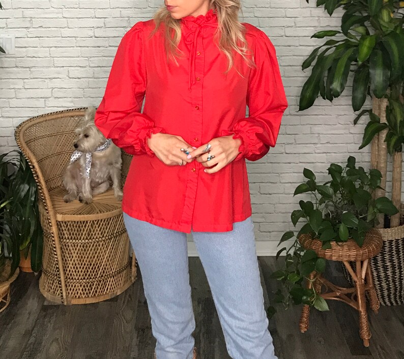 May include: A red long-sleeved blouse with a ruffled collar and button-up front. The blouse has puffed sleeves with ruffles at the cuffs.