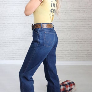 May include: A woman wearing a yellow t-shirt with the text "Doin' it in the canyons" and blue denim jeans with a brown belt. She is standing in front of a white brick wall.