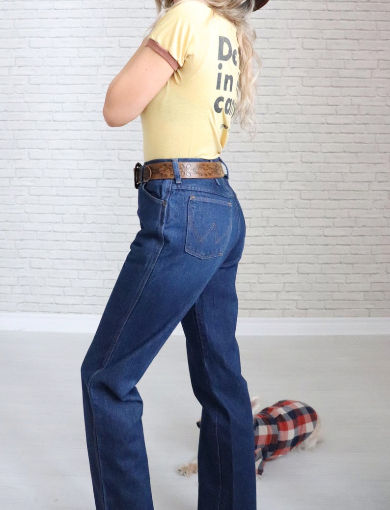 May include: A woman wearing a yellow t-shirt with the text "Doin' it in the canyons" and blue denim jeans with a brown belt. She is standing in front of a white brick wall.