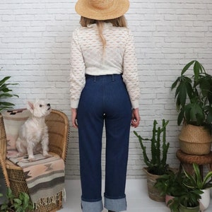 K&ouml;nnte beinhalten: Eine Frau tr&auml;gt einen wei&szlig;en H&auml;kelpullover, eine blaue Jeans mit umgeschlagenen S&auml;umen und einen Strohhut. Sie steht vor einer wei&szlig;en Backsteinwand mit Pflanzen in T&ouml;pfen.