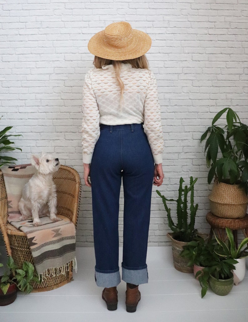 K&ouml;nnte beinhalten: Eine Frau tr&auml;gt einen wei&szlig;en H&auml;kelpullover, eine blaue Jeans mit umgeschlagenen S&auml;umen und einen Strohhut. Sie steht vor einer wei&szlig;en Backsteinwand mit Pflanzen in T&ouml;pfen.