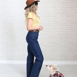 May include: A woman wearing a yellow and brown striped shirt, blue jeans, and a brown cowboy hat stands next to a small white dog wearing a red and black checked shirt.