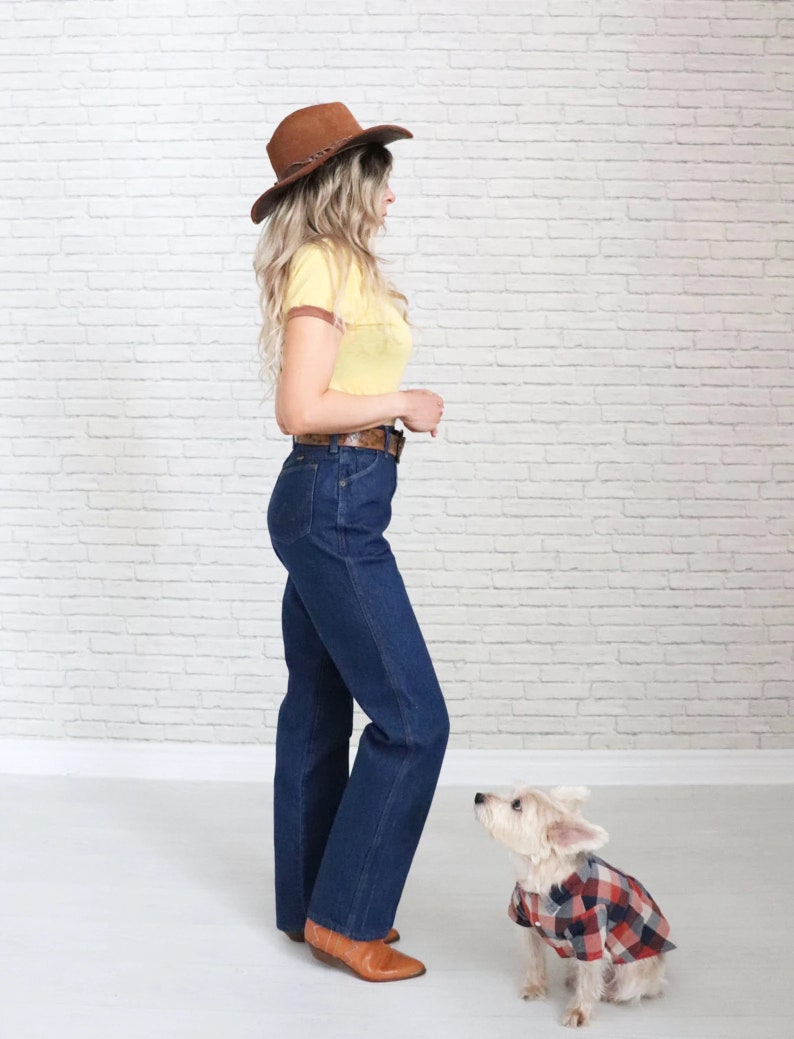 May include: A woman wearing a yellow and brown striped shirt, blue jeans, and a brown cowboy hat stands next to a small white dog wearing a red and black checked shirt.
