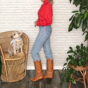 May include: A woman wearing a red ruffled blouse, light blue jeans, and brown cowboy boots. She is standing next to a wicker chair with a small white dog sitting on it.