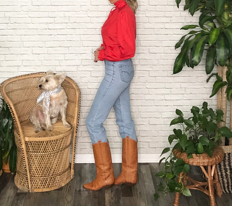 May include: A woman wearing a red ruffled blouse, light blue jeans, and brown cowboy boots. She is standing next to a wicker chair with a small white dog sitting on it.