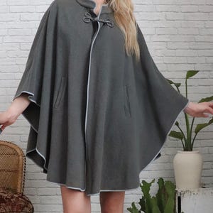 May include: A grey wool cape with a high collar and a decorative rope closure. The cape has a light grey trim and two pockets.