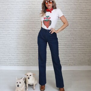 May include: A woman wearing a white t-shirt with a red strawberry graphic and the word "POP" on it. She is also wearing blue jeans, brown boots, and sunglasses. She has a red bandana around her neck. Two small white dogs are sitting in front of her.