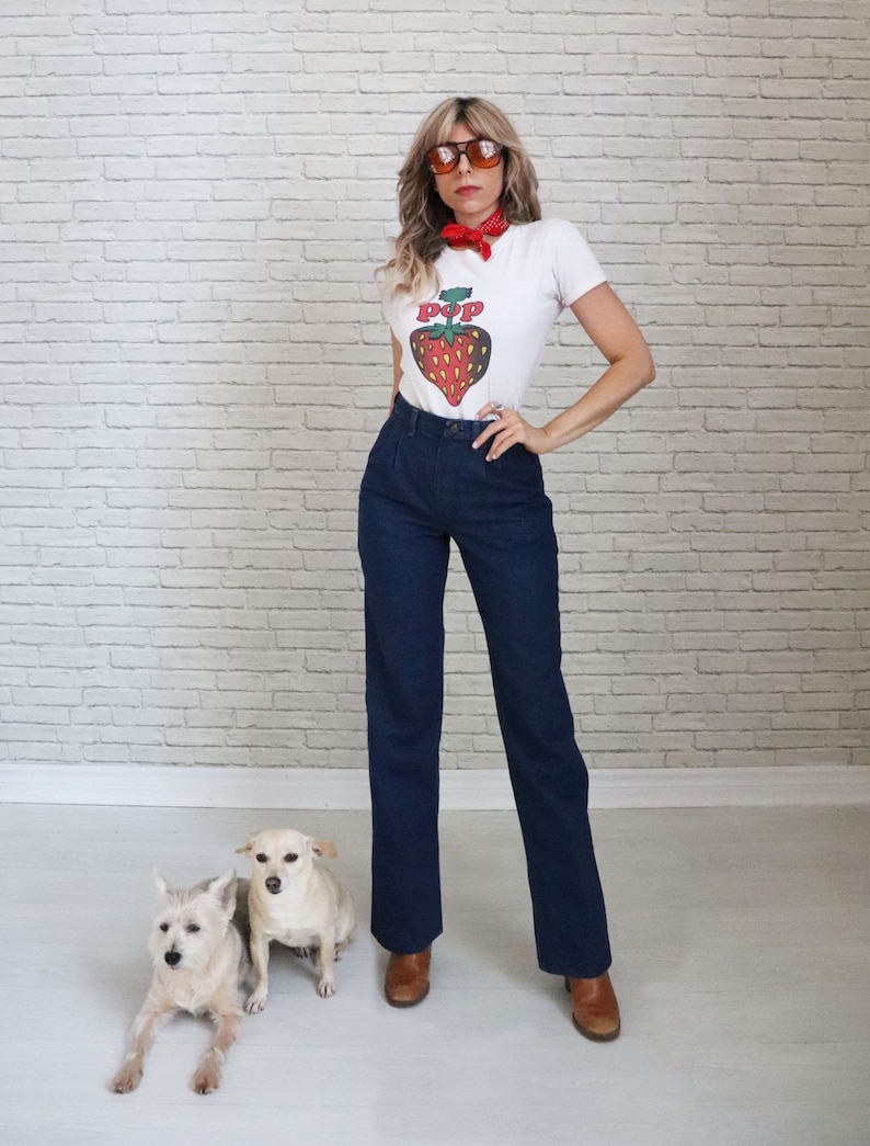 May include: A woman wearing a white t-shirt with a red strawberry graphic and the word "POP" on it. She is also wearing blue jeans, brown boots, and sunglasses. She has a red bandana around her neck. Two small white dogs are sitting in front of her.