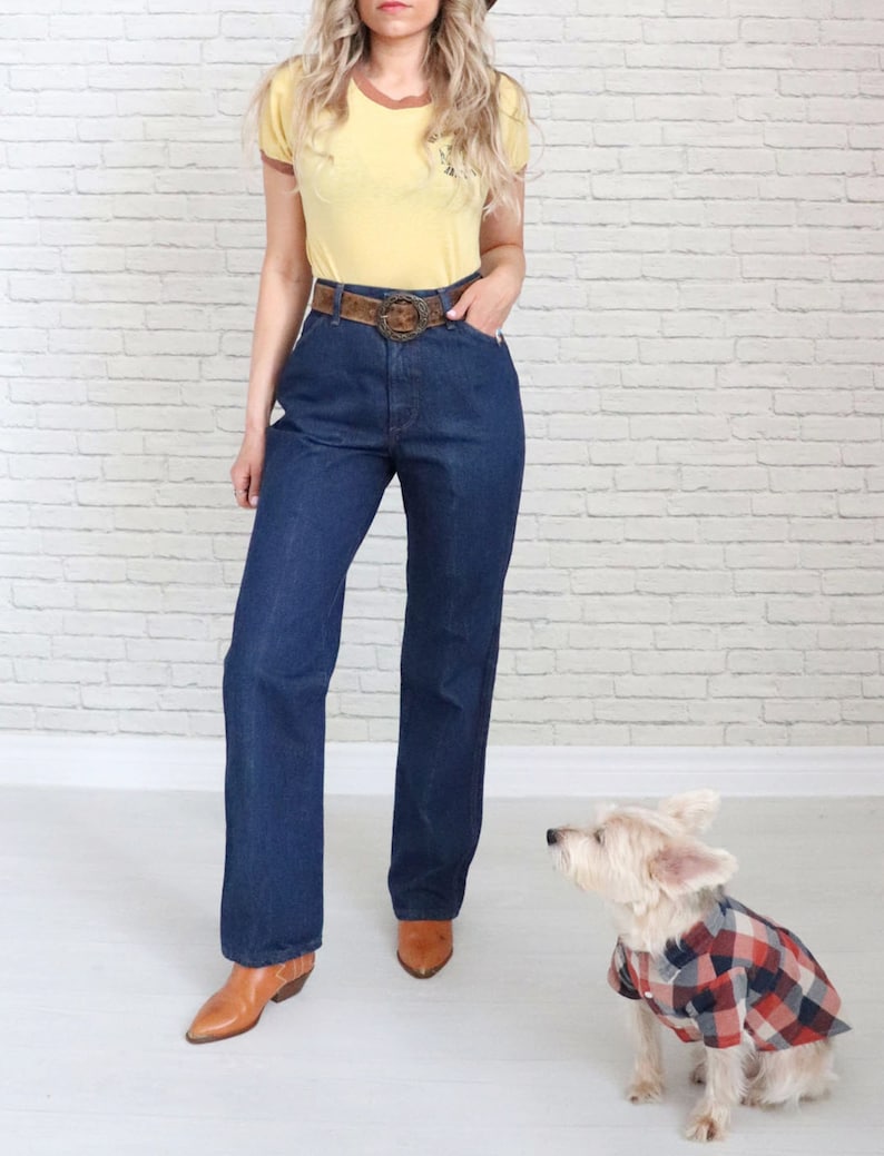 May include: A woman wearing a yellow t-shirt with a brown belt and dark blue jeans. She is wearing brown cowboy boots. A small white dog wearing a red and black checked shirt is sitting in front of her.