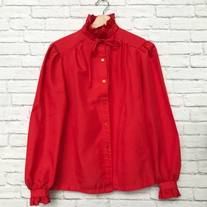May include: A red long-sleeved blouse with a ruffled collar and a bow tie. The blouse has a button-up front and long sleeves with ruffled cuffs.