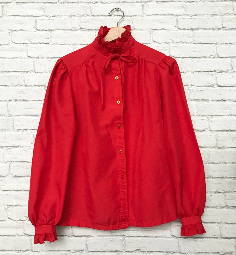 May include: A red long-sleeved blouse with a ruffled collar and a bow tie. The blouse has a button-up front and long sleeves with ruffled cuffs.