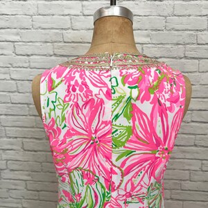 May include: A sleeveless dress featuring a vibrant floral pattern in shades of hot pink, white, and lime green. The neckline is accented with a decorative gold metallic trim. The garment is displayed on a mannequin and includes a center back zipper.