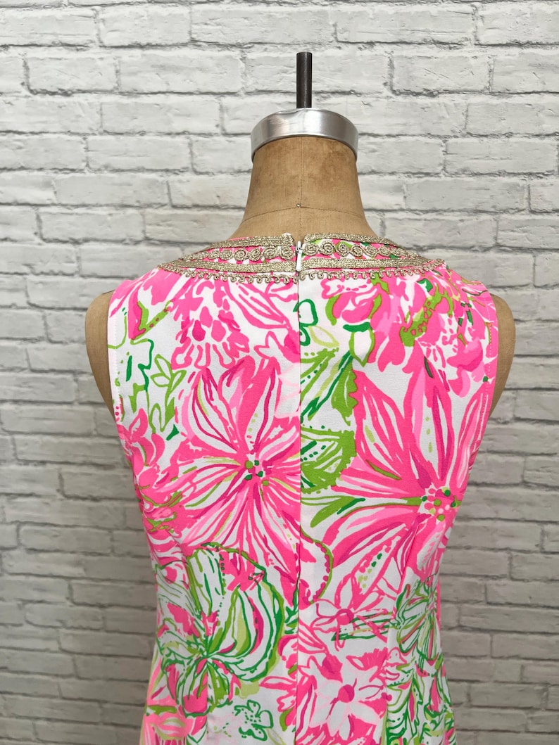 May include: A sleeveless dress featuring a vibrant floral pattern in shades of hot pink, white, and lime green. The neckline is accented with a decorative gold metallic trim. The garment is displayed on a mannequin and includes a center back zipper.