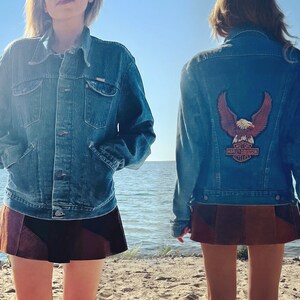 May include: Denim jacket with a Harley-Davidson eagle patch on the back. The jacket is a medium wash blue with a button-up front and pockets. The model is wearing a brown and black skirt.