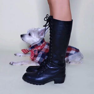 May include: Black knee-high lace-up boots with a small heel. A white dog wearing a red and blue plaid bandana and shirt is lying down next to the boot. The boots have a round toe and a thick sole.