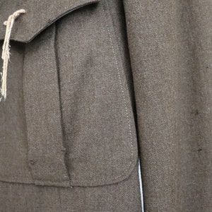 Pu&ograve; includere: Primo piano di una giacca di lana marrone con due tasche anteriori e un risvolto abbottonato. La giacca ha un aspetto strutturato e un cordino colorato chiaro attaccato alla tasca. La giacca &egrave; in stile vintage.