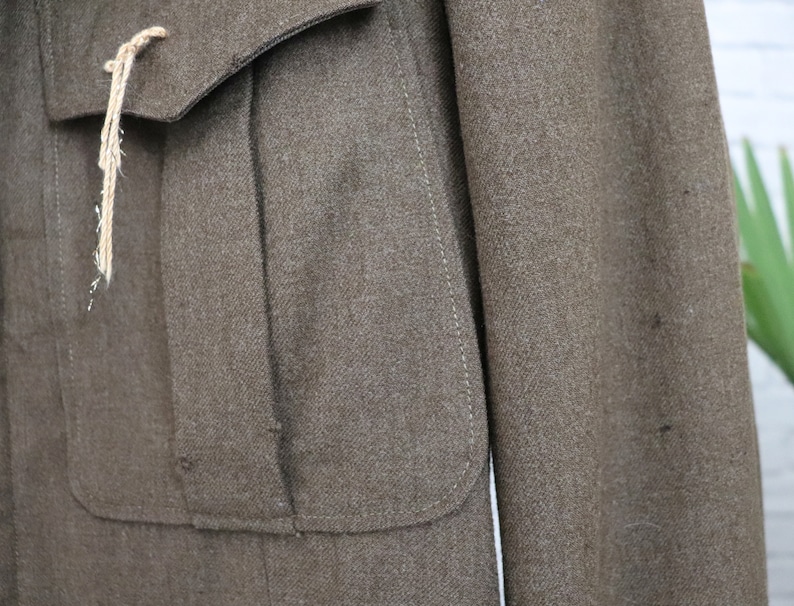 Pu&ograve; includere: Primo piano di una giacca di lana marrone con due tasche anteriori e un risvolto abbottonato. La giacca ha un aspetto strutturato e un cordino colorato chiaro attaccato alla tasca. La giacca &egrave; in stile vintage.