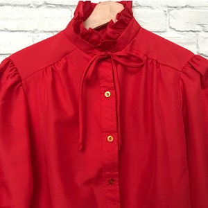 May include: A red long-sleeved blouse with a ruffled collar and a bow tie. The blouse has a button-up front and gold buttons.