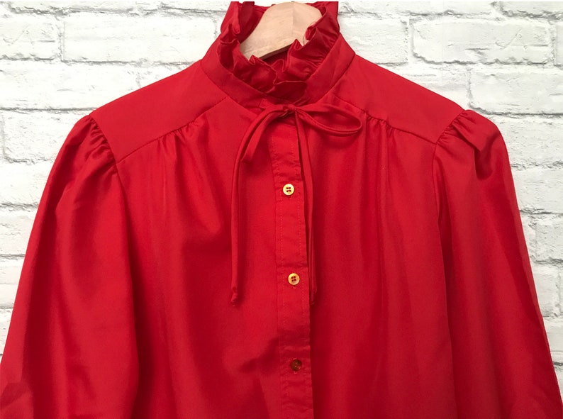 May include: A red long-sleeved blouse with a ruffled collar and a bow tie. The blouse has a button-up front and gold buttons.