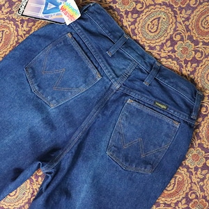 May include: A pair of dark blue denim jeans with a back pocket featuring the Wrangler logo and a stitched 'W' design. The jeans have a high waist and a straight leg.