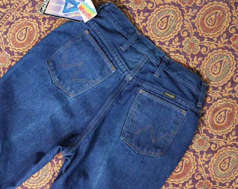 May include: A pair of dark blue denim jeans with a back pocket featuring the Wrangler logo and a stitched 'W' design. The jeans have a high waist and a straight leg.