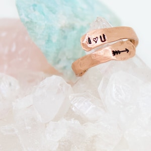 May include: A copper ring with the words "I ♥ U" and an arrow engraved on it. The ring is sitting on a bed of white crystals.