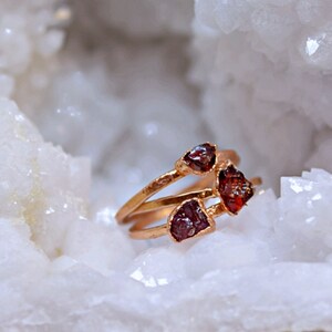 May include: A gold-toned ring with three raw, reddish-brown gemstones. The ring is set against a backdrop of a white, crystalline structure. The gemstones have a rough, natural appearance, adding a unique touch to the jewelry.