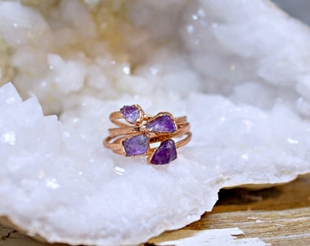 May include: Copper-colored rings with raw amethyst stones. The rings are stacked and displayed on a white, crystalline surface. The amethyst stones are a deep purple color and have a natural, irregular shape. The rings are a unique jewelry item.