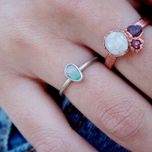 May include: A silver ring with an oval opal stone, displaying iridescent colors of blue, green, and purple. Another ring with a copper band and white and purple stones is also visible.