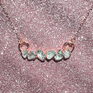 May include: A rose gold necklace featuring a row of six light blue, irregularly shaped gemstones. The gemstones are set in a curved bar, suspended from a delicate chain. The background is a sparkly, pink surface.