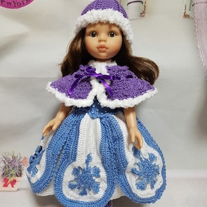May include: A doll wearing a handmade crochet outfit. The outfit includes a purple hat with white trim, a matching cape, and a blue and white dress with snowflake designs. The doll has brown hair and is standing.