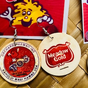 Puede incluir: Un par de pendientes con diseños vintage de tapas de POG (Passion Orange Guava). Un pendiente muestra el logotipo de "Orchards Hawaii Inc", el otro el logotipo de "Meadow Gold". Los pendientes están suspendidos de ganchos plateados.