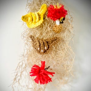 May include: A whimsical, handmade wall hanging featuring a face-like design. The artwork incorporates yellow crochet, a red pom-pom, and a brown crocheted mouth with pearl accents. The background is composed of dried, light brown plant fibers.