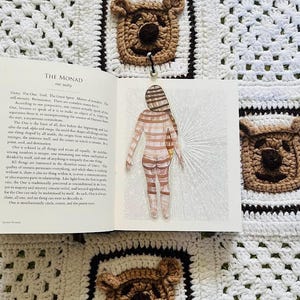 May include: An open book titled "THE MONAD" with a drawing of a figure in striped clothing. The book rests on a white crocheted blanket with brown bear-shaped appliqués. The blanket has a black border.