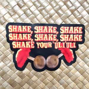 Könnte beinhalten: Ein schwarzer Aufkleber mit dem Text "SHAKE, SHAKE, SHAKE SHAKE, SHAKE, SHAKE SHAKE YOUR ULI ULI" in Rot und Weiß. Der Aufkleber hat ein Design von zwei braunen Objekten mit roten Akzenten. Der Hintergrund ist ein gewebtes Muster.